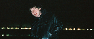 Eine Person in schwarzer Lederjacke steht nachts draußen. Hintergrund zeigt verschwommene Lichter.