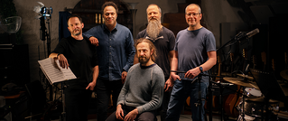 Fünf Männer in einem Musikstudio, vier stehen, einer sitzt. Instrumente und Mikrophon sind im Hintergrund. Atmosphäre ist entspannt.