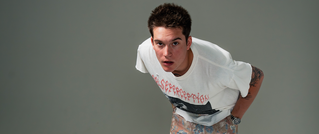 Ein Mann in einem weißen T-Shirt beugt sich nach vorne. Der Hintergrund ist neutral grau.