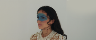 Frau mit blauem Augen-Make-up und einem perlenbesetzten Oberteil vor hellem Hintergrund, schaut nach links.