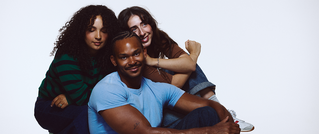 Drei Personen lächeln, sitzen nah zusammen. Ein Mann in blauem T-Shirt vorne, zwei Frauen in entspannter Pose dahinter vor weißem Hintergrund.
