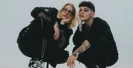 Zwei Personen in dunkler Kleidung hocken nebeinander. Eine trägt eine Brille, beide haben Tattoos und schauen in die Kamera.