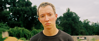 Eine Person mit kurzem Haar schaut ernst in die Kamera. Sie trägt ein dunkles Shirt. Der Hintergrund zeigt Bäume und Himmel.