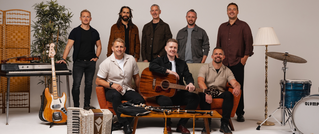 Acht Männer posieren mit Instrumenten, darunter Gitarre, Akkordeon und Schlagzeug, in einem Studio mit Pflanzen und Lampen.