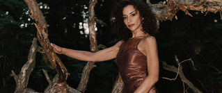 Eine Person in einem braunen Kleid steht im Wald neben einem verwachsenen Baum. Es ist Abend, das Licht ist gedämpft.