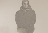 Eine Person in einem pelzartigen Mantel sitzt mit überkreuzten Beinen vor neutralem Hintergrund. Das Bild ist in sepia gehalten.