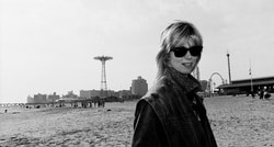 Eine Person mit Sonnenbrille steht am Strand vor der Skyline von Coney Island. Im Hintergrund sind Fahrgeschäfte zu sehen.