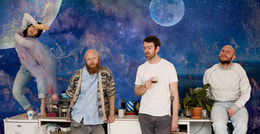 Vier Personen posieren vor einem fantasievollen Hintergrund mit Planeten und Sternen. Einer steht auf dem Tisch und tanzt.