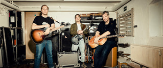 Drei Männer mit Gitarren stehen in einem Proberaum, umgeben von Verstärkern und Instrumentenkoffern.