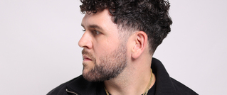 Ein Mann mit lockigem dunklem Haar und Bart schaut zur Seite vor neutralem Hintergrund. Er trägt eine schwarze Jacke und eine Kette.