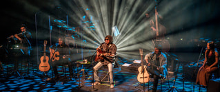 Eine Musikgruppe sitzt auf einer Bühne mit akustischen Gitarren. Im Hintergrund sind Strahler, die Lichtstrahlen erzeugen. Die Bühne ist atmosphärisch beleuchtet, und das Ensemble scheint in einem Konzert zu spielen.