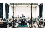 Ein Dirigent steht vor einem Orchester in einem großen Saal. Mikrofone sind aufgestellt, Musiker spielen verschiedene Instrumente.