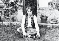 Eine Person sitzt auf dem Rasen vor einem Baum im Garten, umgeben von Pflanzen in Töpfen. Das Bild ist schwarz-weiß.