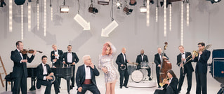 In einem hellen Studio stehen und sitzen Musiker einer Big Band, gekleidet in formellen Anzügen. Ein Pianist spielt Klavier, während Geiger, Bläser und ein Schlagzeuger sie umgeben. Eine zentrale Figur tanzt oder posiert in einem glitzernden Kleid. Über ihnen hängen Leuchten von der Decke.