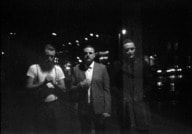 Schwarz-weiß Foto von drei Personen auf einer nächtlichen Straße, verschwommen und unter Beleuchtung von Straßenlaternen.
