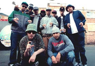 Eine Gruppe von elf Personen posiert draußen, viele tragen Hüte und machen Handgesten. Sie stehen vor einem Backsteingebäude.