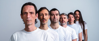 Sechs Männer in weißen T-Shirts stehen hintereinander in diagonaler Linie, jeweils einer nach dem anderen, vor einem grauen Hintergrund.