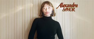 Eine Person in einem schwarzen Rollkragenpullover steht vor einer gestreiften Wand. Oben rechts steht in roter Schrift 'Alexandra Savior'.
