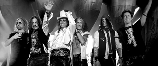 Eine Gruppe von sechs Menschen steht auf einer Bühne, einer trägt einen weißen Hut und winkt, alle wirken fröhlich. Schwarz-weiß Bild.