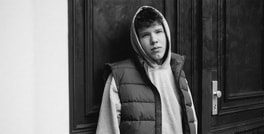 Junger Mann mit Kapuzenpullover und gesteppter Weste lehnt an einer dunklen Tür, Blick nachdenklich in die Ferne gerichtet.
