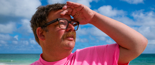 Ein Mann mit Brille und Schnurrbart blickt in die Ferne. Er trägt ein rosa T-Shirt, steht am Strand unter blauem Himmel mit Wolken.