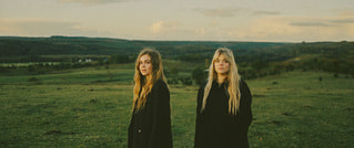 Zwei Frauen mit langen Haaren stehen in einer weiten, grünen Landschaft unter bewölktem Himmel, beide in dunkler Kleidung.