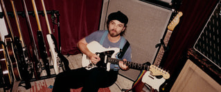 Eine Person sitzt auf einem Teppich, spielt E-Gitarre. Umgeben von mehreren Gitarren und einem Verstärker, trägt sie eine Mütze.