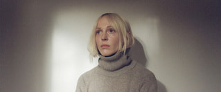 Person mit blondem Haar in hellem Rollkragenpullover vor einem neutralen, schattenhaften Hintergrund. Blick nachdenklich in die Ferne.