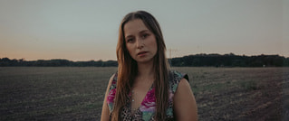 Eine Frau mit langen Haaren steht auf einem Feld bei Sonnenuntergang. Sie trägt ein gemustertes Oberteil und blickt ernst in die Kamera.