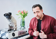 Ein Mann in roter Jacke steht neben einem Keyboard mit einem Mops darauf; dahinter ein Blumenstrauß in einer Vase.