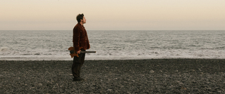 Eine Person steht mit einer Gitarre am steinigen Strand und blickt ins Meer. Der Himmel ist klar und der Horizont ruhig.