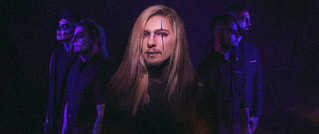 Eine Band posiert mit einem Mann im Vordergrund, bemaltem Gesicht und langen Haaren. Andere Mitglieder sind dunkel gekleidet im Hintergrund.