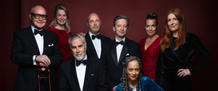 Acht Personen in festlicher Kleidung, fünf Männer in Anzügen und drei Frauen in eleganten Kleidern, posieren vor einem roten Hintergrund.