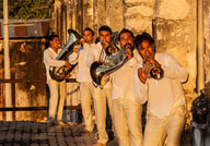 Eine Gruppe von fünf Musikern in weißer Kleidung spielt Blechblasinstrumente vor einer alten Steinwand im Sonnenuntergang.