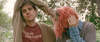 Zwei Personen im Freien, eine mit braunem Shirt, die andere mit rosa Haaren, das Gesicht teilweise mit der Hand verdeckt.
