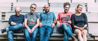 Fünf Personen sitzen lachend auf Bänken, lässig gekleidet in Jeans und T-Shirts, in freundlicher, entspannter Atmosphäre.