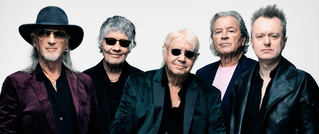 Fünf Männer in dunkler Kleidung posieren vor neutralem Hintergrund. Einige tragen Sonnenbrillen, einer einen Hut.