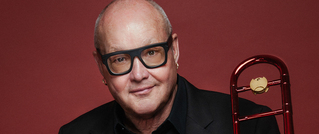 Ein lächelnder Mann mit Brille hält eine rote Posaune vor einem dunkelroten Hintergrund.