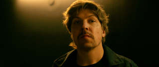 Ein Mann mit Bart und leichtem Lächeln blickt in eine Kamera. Der Hintergrund ist dunkel, mit einem Lichtschein oben.