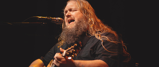 Ein Mann mit langer, blonder Mähne und Bart singt leidenschaftlich in ein Mikrofon und spielt Akustikgitarre auf einer Bühne.