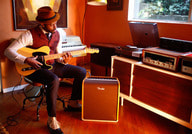 Mann spielt E-Gitarre in einem gemütlichen Raum mit Retro-Musikequipment und einem Verstärker. Warmes Licht von einem Fenster.