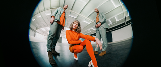 Drei Personen posieren in einem leeren Raum mit moderner Architektur. Eine Person kniet in leuchtend orangefarbenem Outfit.
