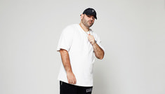 Ein Mann in weißem T-Shirt und schwarzer Kappe steht vor einem neutralen Hintergrund und blickt in die Kamera.