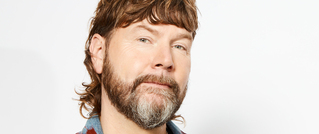 Ein Mann mit braunem, welligem Haar und Bart schaut in die Kamera. Er trägt ein kariertes Hemd. Der Hintergrund ist weiß.
