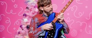 Mann mit Gitarre vor einem pinken Hintergrund mit einem glitzernden Weihnachtsbaum, bekleidet mit einer bunten Jacke.