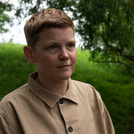Eine Person mit kurzem Haar steht im Freien vor einer grünen, bewaldeten Kulisse und trägt ein beiges Hemd.