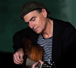 Ein Mann mit Mütze spielt konzentriert Gitarre, trägt ein gestreiftes Shirt und einen dunklen Blazer. Hintergrund dunkel.