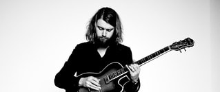 Eine Person mit schulterlangem Haar spielt eine E-Gitarre vor einem weißen Hintergrund. Sie trägt dunkle Kleidung.