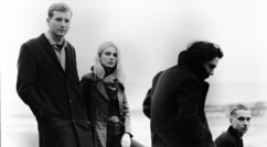 Vier Personen in Mänteln stehen und sitzen am Strand, blicken ernst in schwarz-weißer Fotografie. Hintergrund verschwommen.