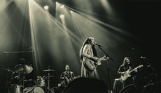 Eine Band spielt auf einer Bühne in dramatischer Beleuchtung. Die Sängerin steht mit Gitarre im Vordergrund, Musiker im Hintergrund.